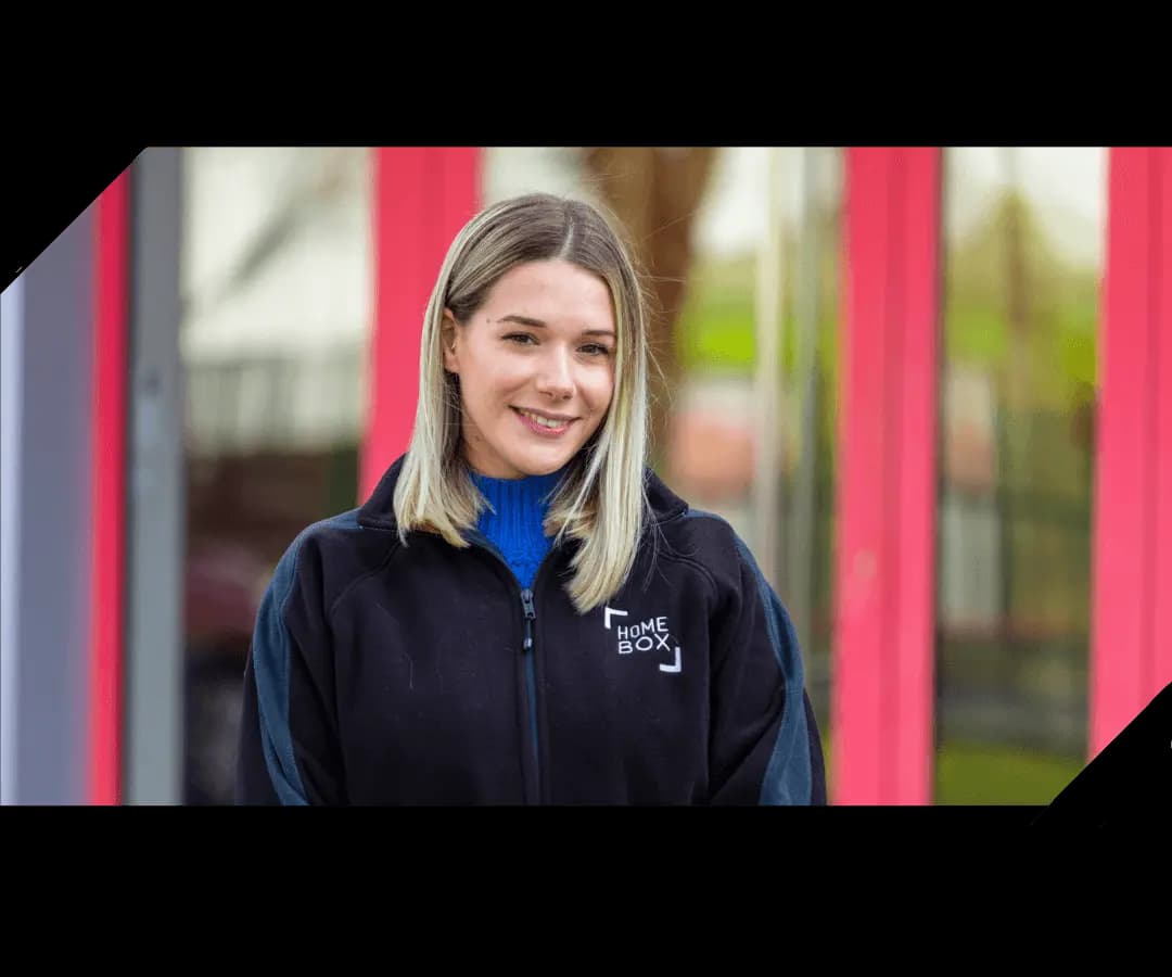 Smiling young woman wearing a HOMEBOX branded jacket in front of a storage centre facade