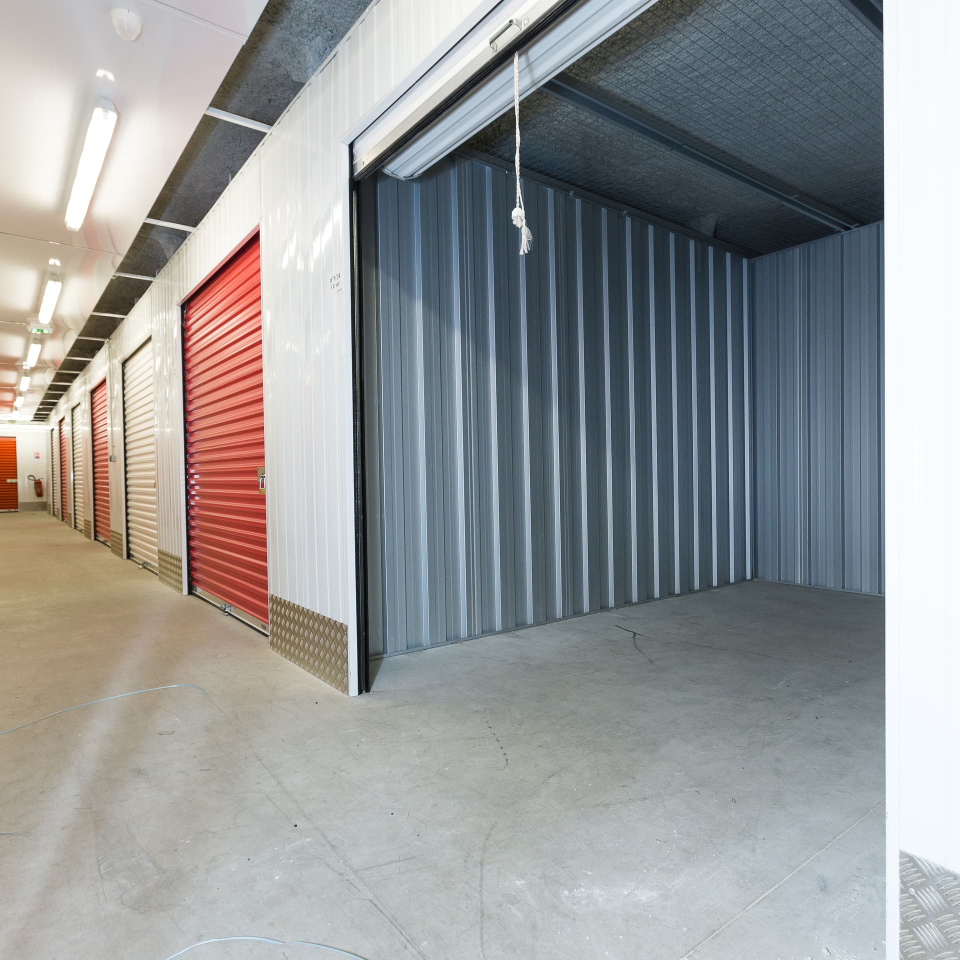 Trastero vacío con suelo de hormigón y pasillo de puertas rojas iluminado del centro HOMEBOX Humanes