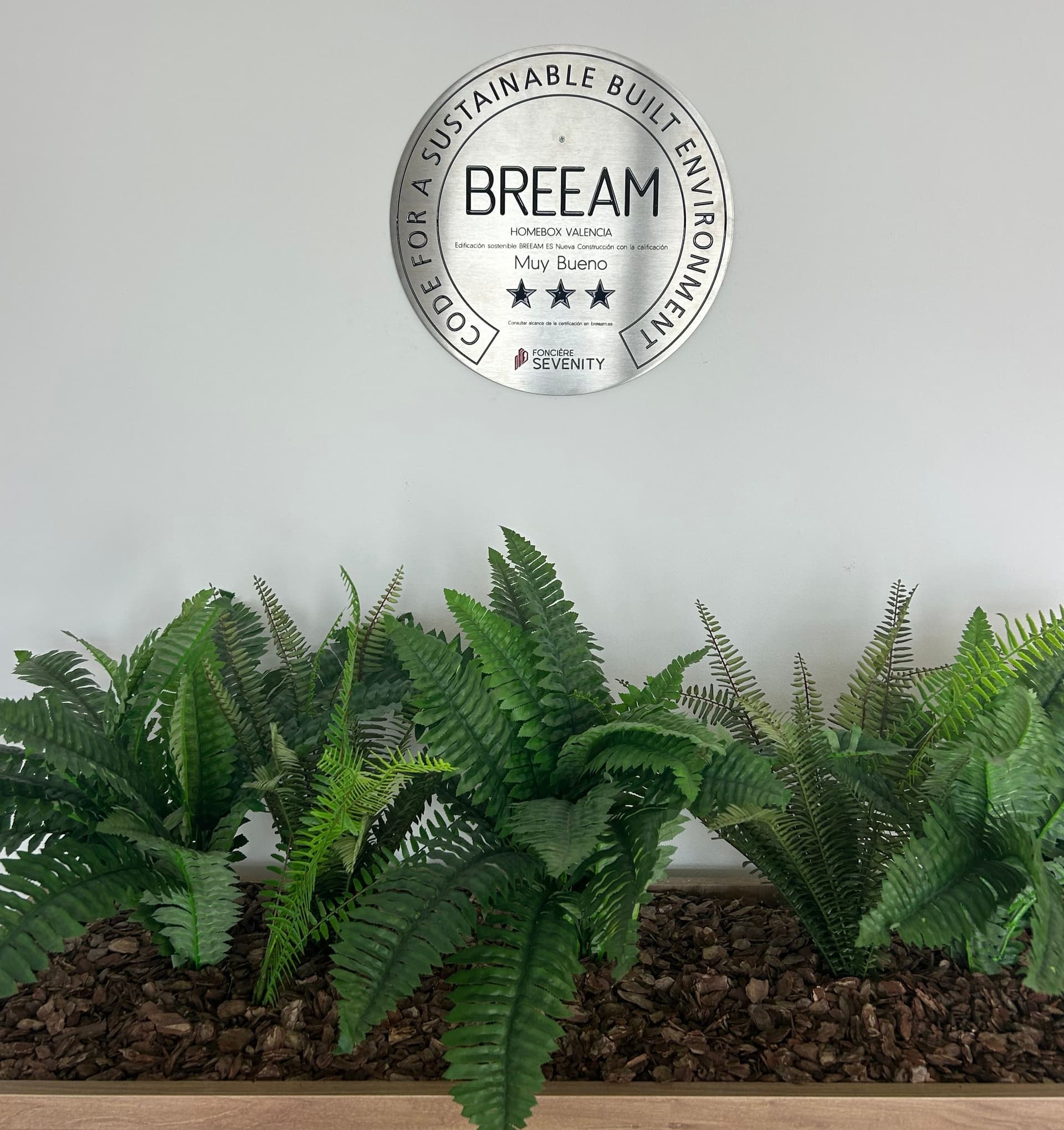 Placa de certificación BREEAM sobre pared blanca con helechos verdes decorativos del centro HOMEBOX Valencia